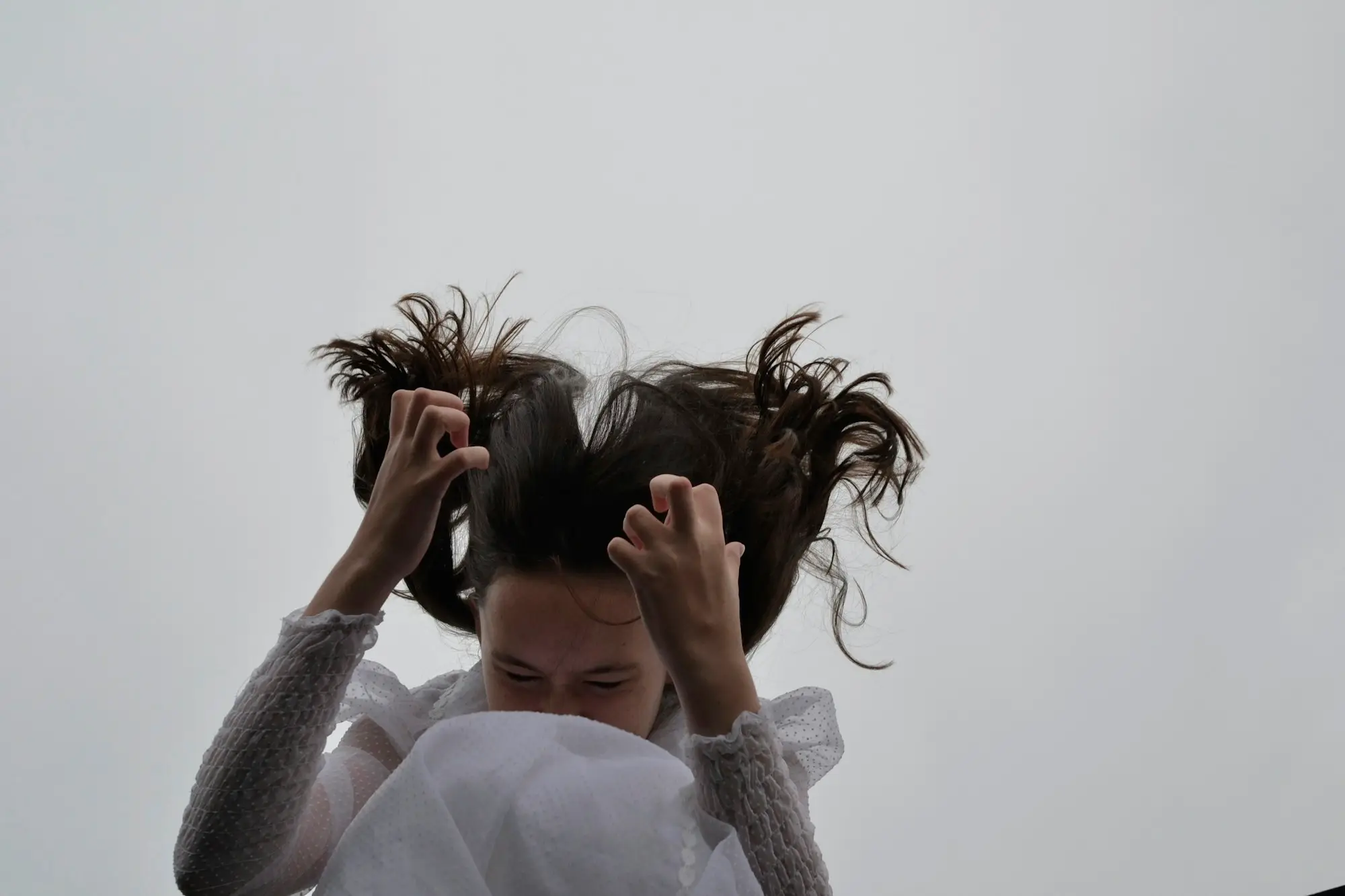 A woman with long dark hair clutches at it with both hands against an overcast neutral background.