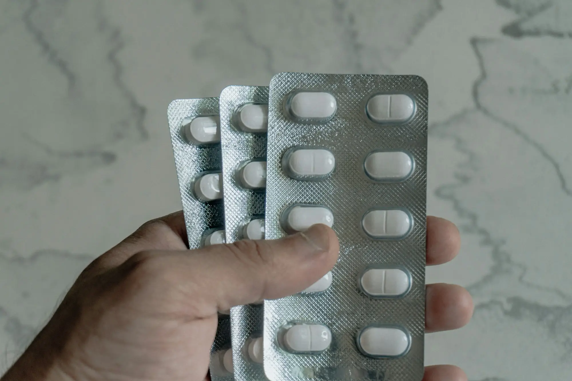A hand holding three unlabelled blister packs of white tablets against a marble background.