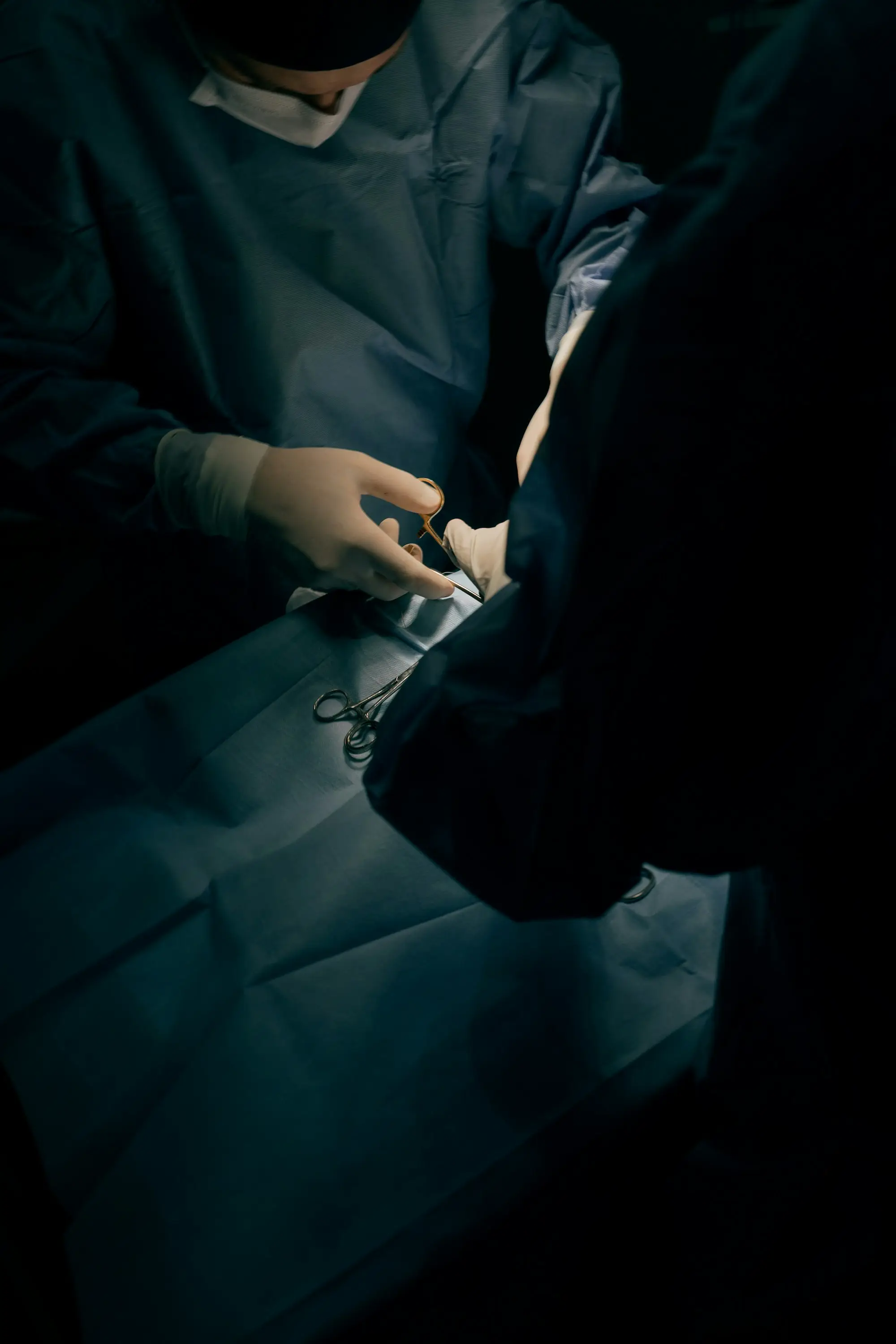 A surgeon in dark surgical attire and gloves works with instruments under directional clinic light.