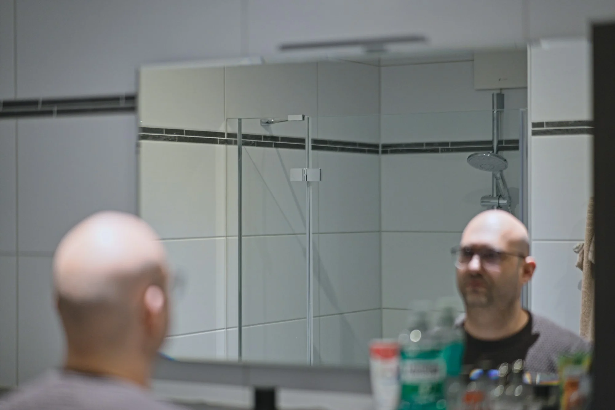 A bald man in glasses looking at himself in a bathroom mirror.