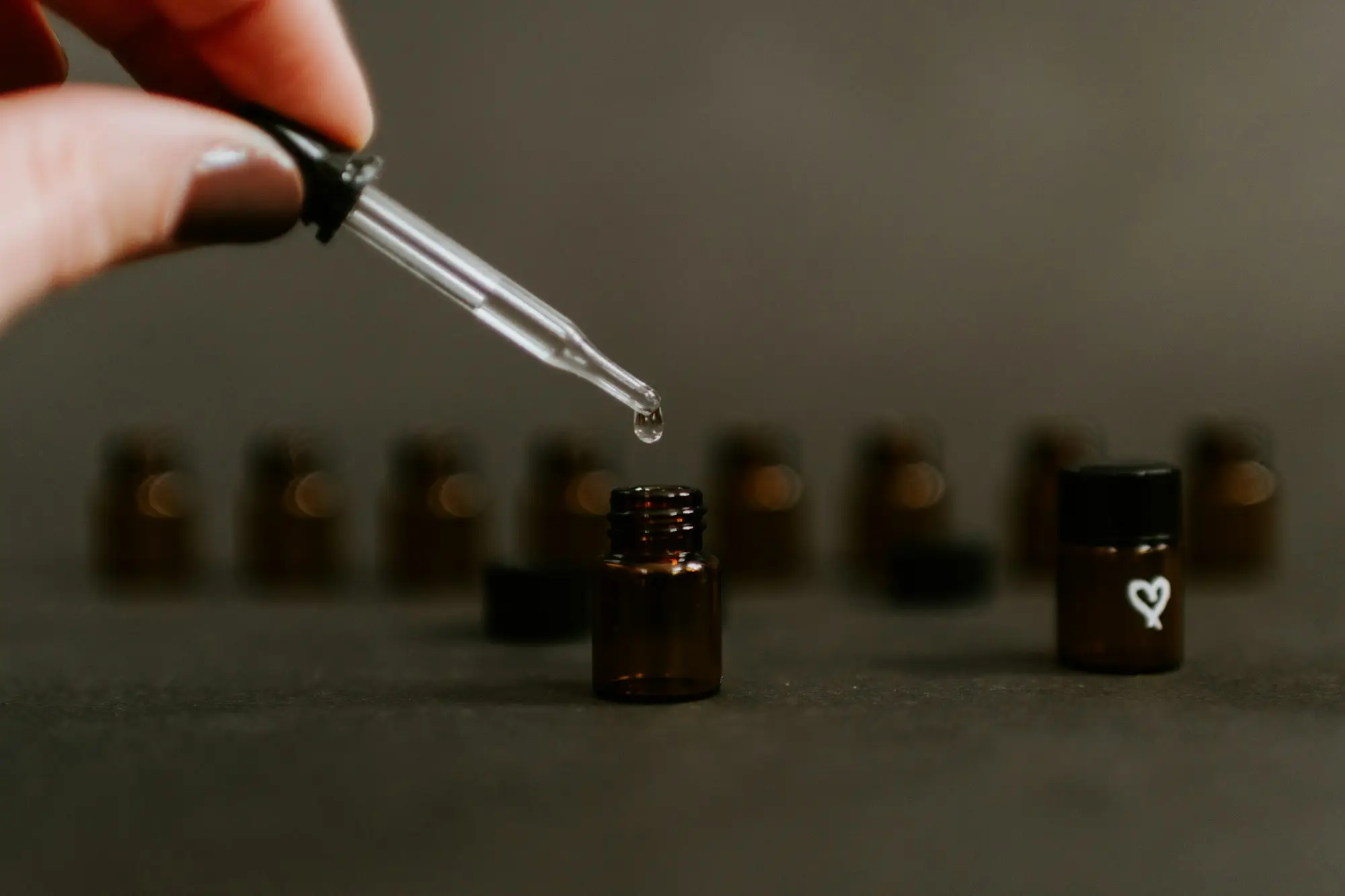 A hand holds a glass dropper above a row of small amber bottles in moody editorial light.