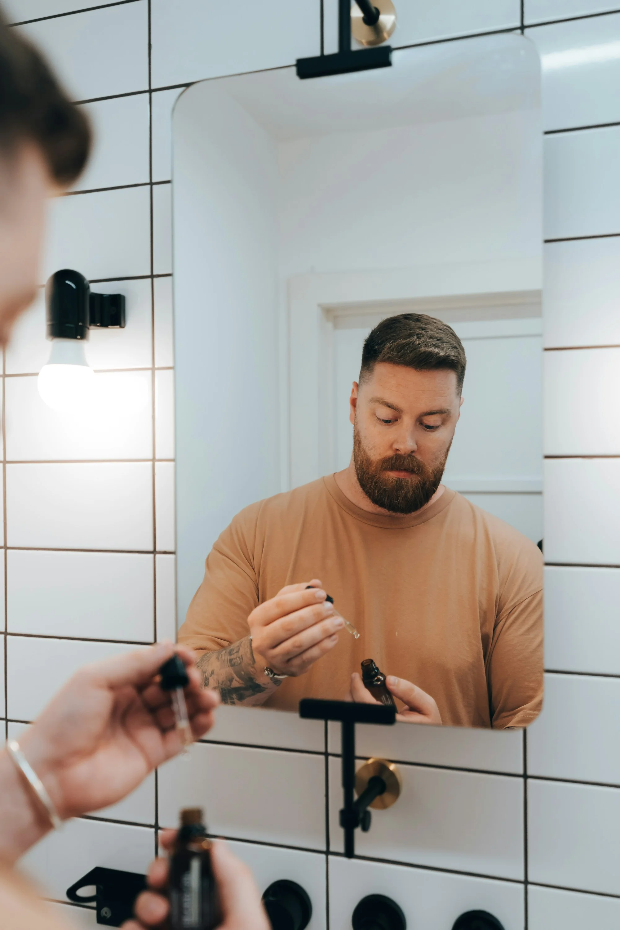 A bearded man in his 40s applying a hair serum at his bathroom mirror.