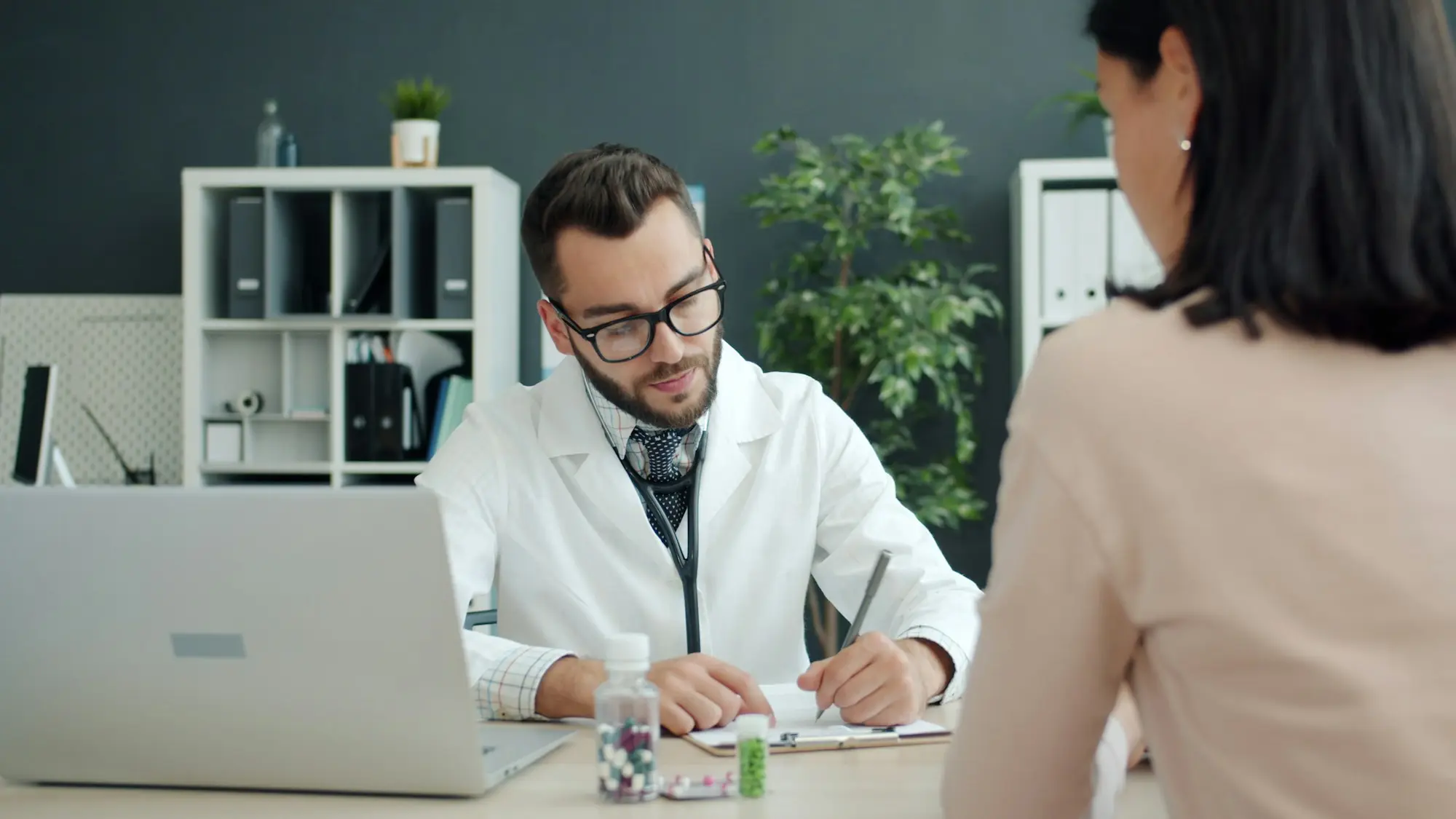A doctor in a white coat with a stethoscope reviews notes with a patient across a desk in a modern clinical office.